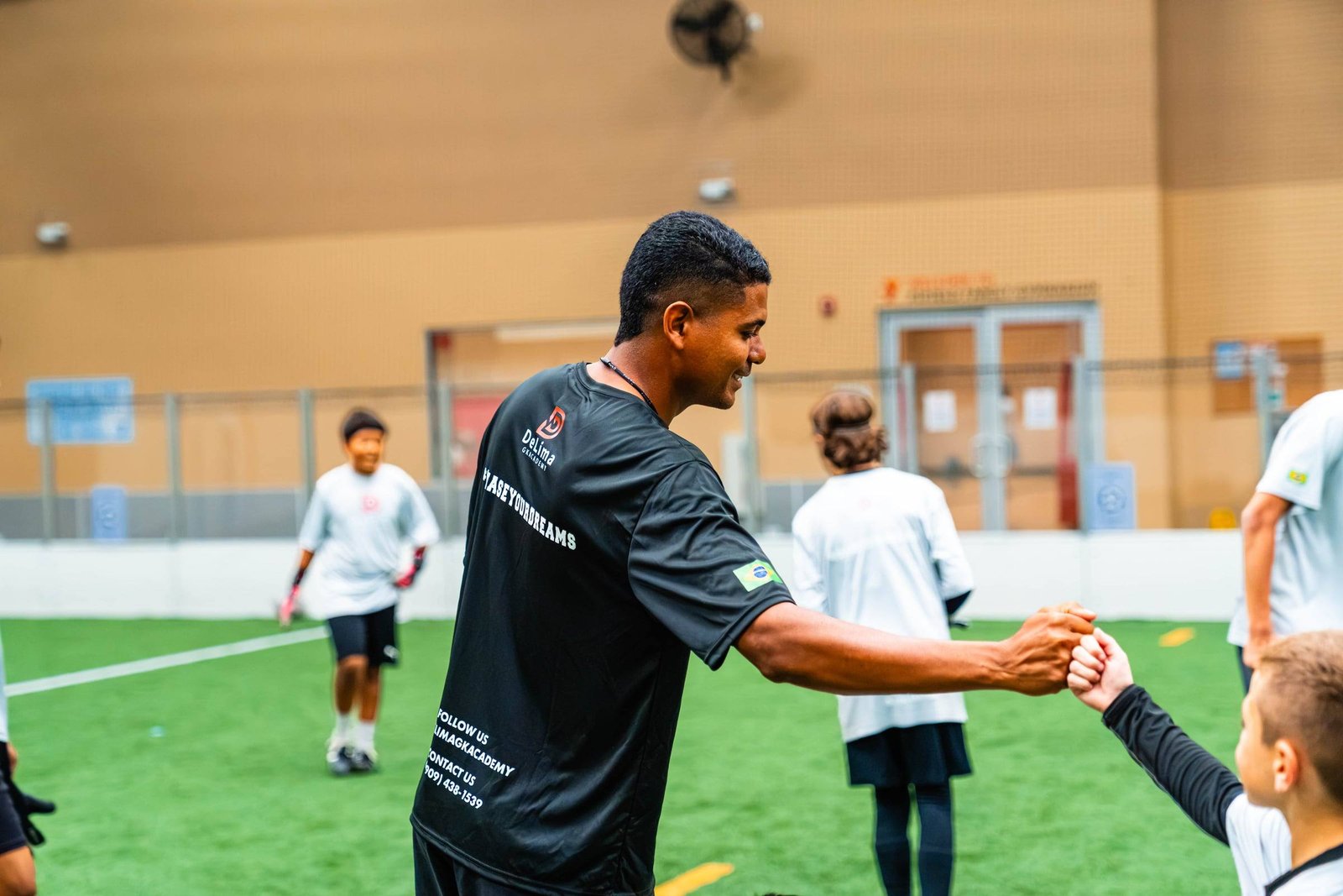 Coach Claysson De Lima smiling and bumping fists with a young goalkeeper during an indoor session in Upland